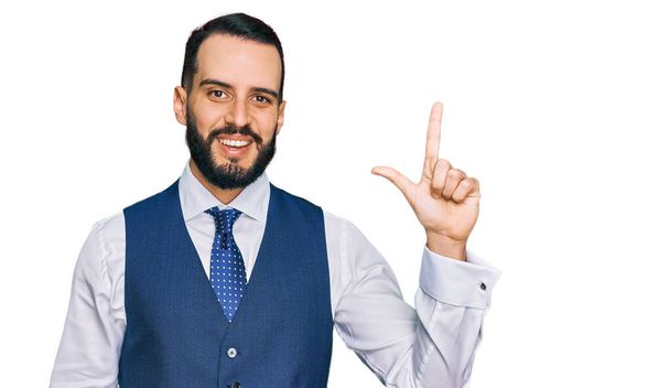 Молодой человек с бородой в жилете для бизнеса показывает и показывает пальцами номер два, улыбаясь уверенно и счастливо.  - Фото, изображение