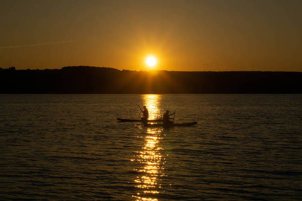 two surfers sitting and row on standup paddleboards against the backdrop of the setting su - Photo, image