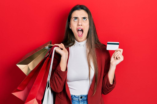 jong brunette tiener holding shopping tassen en credit card boos en gek geschreeuw gefrustreerd en furieus, schreeuwen met woede omhoog te kijken.  - Foto, afbeelding