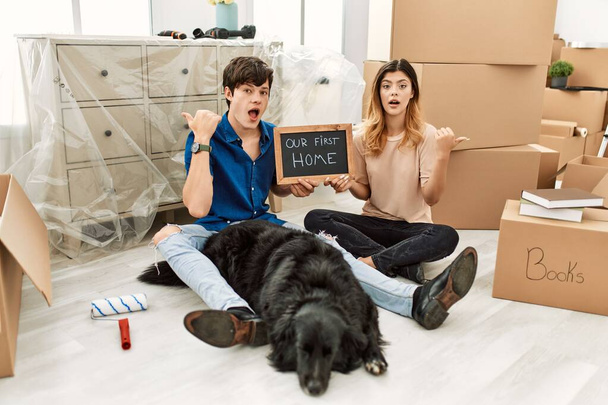 Young caucasian couple with dog holding our first home blackboard at new house surprised pointing with hand finger to the side, open mouth amazed expression.  - Photo, Image