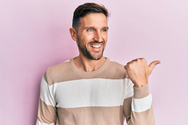 Красивый мужчина с бородой в повседневной одежде указывает пальцем вверх в сторону улыбается счастливо с открытым ртом  - Фото, изображение