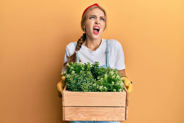 Beautiful blonde caucasian woman holding wooden plant pot angry and mad screaming frustrated and furious, shouting with anger looking up.  - Fotoğraf, Görsel