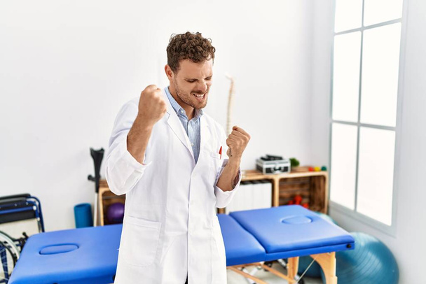 Pohledný mladý muž pracující na klinice pro zotavení bolesti velmi šťastný a vzrušený dělá vítězný gesto se zdviženýma rukama, usmívá se a křičí po úspěchu. koncepce oslav.  - Fotografie, Obrázek