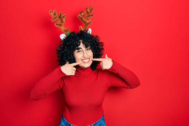 Młoda kobieta na Bliskim Wschodzie nosi słodkie rogi reniferów Boże Narodzenie uśmiechając wesoły pokaz i wskazując palcami zęby i usta. Koncepcja zdrowia zębów.  - Zdjęcie, obraz