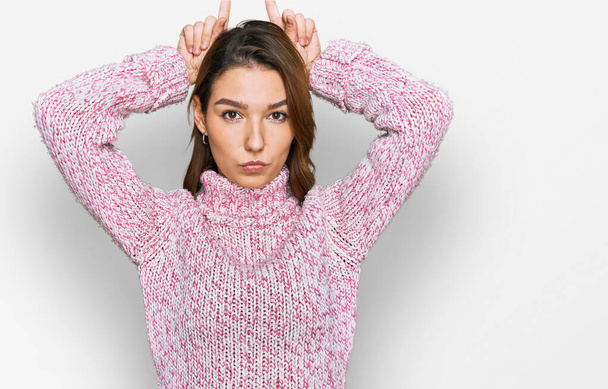 Joven chica caucásica con suéter de lana de invierno haciendo gesto divertido con el dedo sobre la cabeza como cuernos de toro  - Foto, Imagen