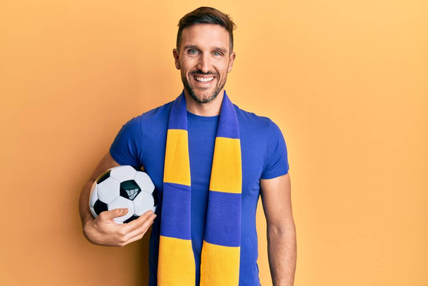 Handsome man with beard football hooligan cheering game holding ball looking positive and happy standing and smiling with a confident smile showing teeth  - Photo, Image