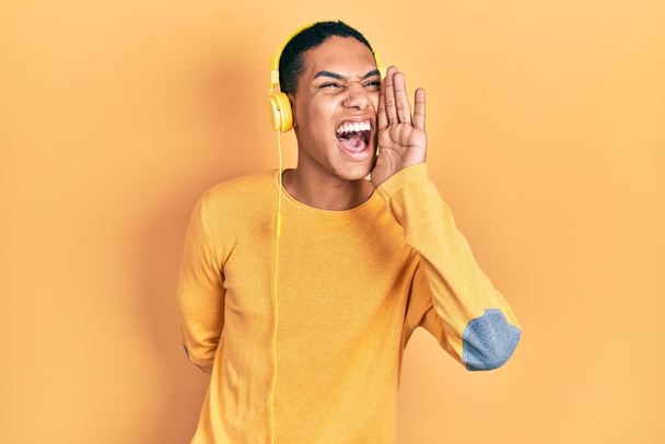 Νεαρός Αφροαμερικάνος που ακούει μουσική με ακουστικά φωνάζοντας και φωνάζοντας δυνατά στο πλάι με το χέρι στο στόμα. έννοια επικοινωνίας.  - Φωτογραφία, εικόνα