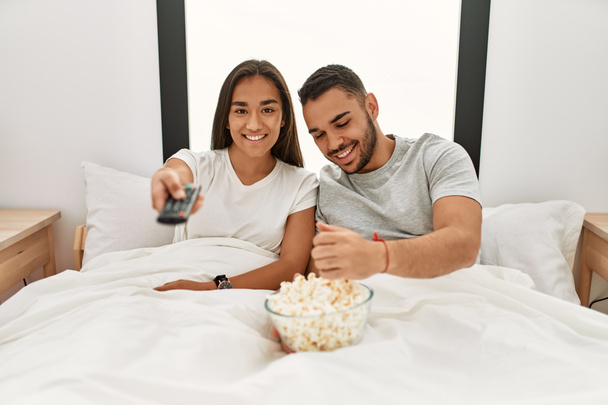 Joven pareja latina viendo películas y comiendo palomitas de maíz acostada en la cama. - Foto, Imagen