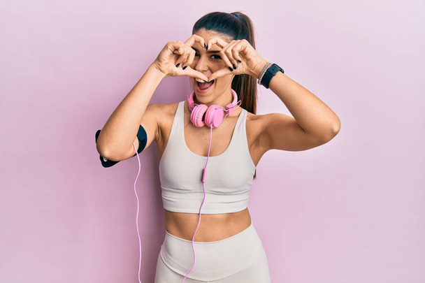 Young hispanic woman wearing gym clothes and using headphones doing heart shape with hand and fingers smiling looking through sign  - Foto, Imagem
