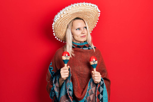 Belle femme blonde caucasienne portant poncho mexicain festif et maracas souriant regardant vers le côté et regardant loin en pensant.  - Photo, image