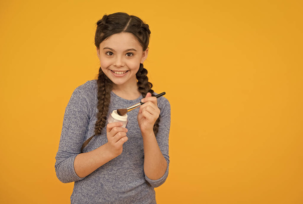 smiling kid girl with stylish braided hair hold loose powder with applicator on yellow background, copy space, skin care - Photo, Image
