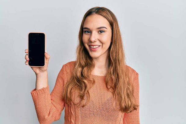 jong blond vrouw holding smartphone tonen scherm kijken positief en gelukkig staan en glimlachen met een zelfverzekerde glimlach tonen tanden  - Foto, afbeelding