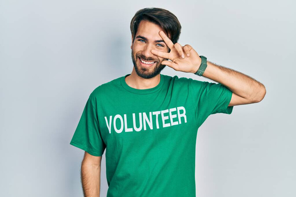 Junger hispanischer Mann im freiwilligen T-Shirt, der das Friedenssymbol mit den Fingern über dem Gesicht trägt, lächelt fröhlich und zeigt den Sieg  - Foto, Bild