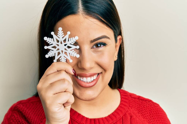 Όμορφη μελαχρινή γυναίκα κρατώντας νιφάδα χιονιού πάνω από τα μάτια αναζητούν θετική και ευτυχισμένη στέκεται και χαμογελά με αυτοπεποίθηση χαμόγελο δείχνει τα δόντια  - Φωτογραφία, εικόνα
