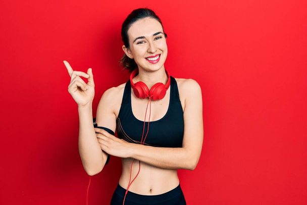 Νεαρό ισπανόφωνο κορίτσι που φοράει ρούχα γυμναστικής και χρησιμοποιεί ακουστικά με ένα μεγάλο χαμόγελο στο πρόσωπο, δείχνοντας με το χέρι και το δάχτυλο στο πλάι κοιτάζοντας την κάμερα.  - Φωτογραφία, εικόνα