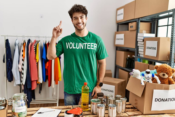 Hispanic man wearing volunteer t shirt at donations stand surprised with an idea or question pointing finger with happy face, number one  - Fotó, kép