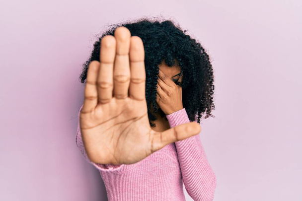 Afroamerykanka z afro włosy noszące zwykłą różową koszulę pokrywającą oczy dłońmi i wykonując stop gest ze smutkiem i wyrazem strachu. wstydliwa i negatywna koncepcja.  - Zdjęcie, obraz