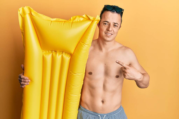 Schöner junger Mann im Badeanzug und mit sommerlichem Schwimmer lächelnd glücklich mit Hand und Finger zeigend  - Foto, Bild