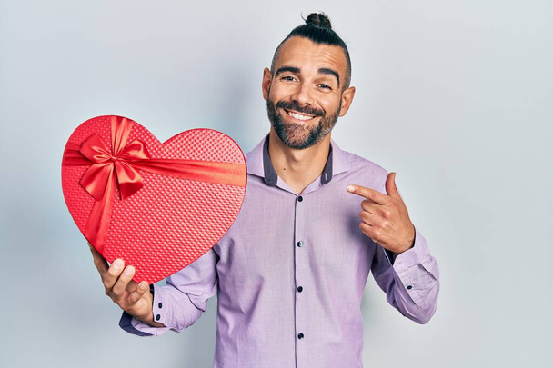 Junger hispanischer Mann mit Valentinstagsgeschenk lächelnd glücklich mit Hand und Finger zeigend  - Foto, Bild