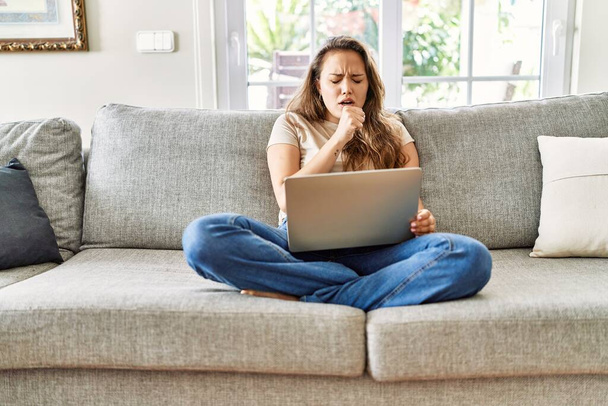 Belle jeune femme brune assise sur le canapé utilisant un ordinateur portable à la maison se sentant mal à l'aise et toussant comme symptôme du froid ou de la bronchite. concept de soins de santé.  - Photo, image