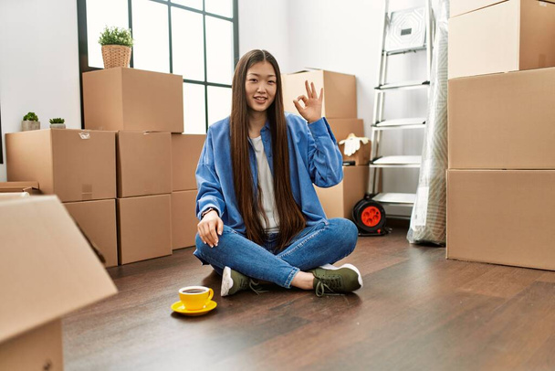 Mladá Číňanka sedící na podlaze v novém domově a usmívající se pozitivně, dělá v pořádku ceduli s rukama a prsty. úspěšný výraz.  - Fotografie, Obrázek