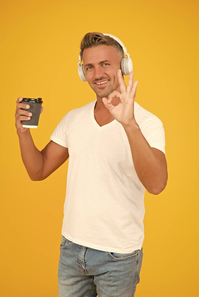 Happy man listen to music showing OK hand gesture for takeaway coffee in disposable cup yellow background, coffee-to-go - Foto, immagini