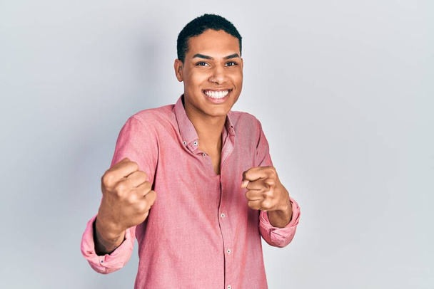若いアフリカ系アメリカ人の男性は、カジュアルな服を着て非常に満足し、腕を上げて勝者のジェスチャーを行う興奮し、笑顔と成功のために叫んでいます。お祝いのコンセプト.  - 写真・画像