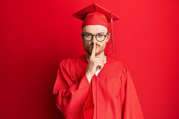 Giovane uomo rossa con cappello rosso graduazione e accappatoio cerimonia chiedendo di essere tranquillo con il dito sulle labbra. silenzio e concetto segreto.  - Foto, immagini