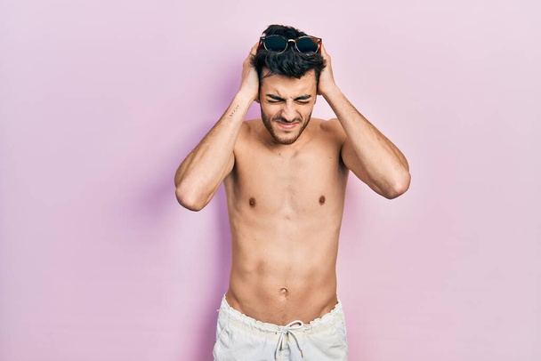 Jonge Latijns-Amerikaanse man draagt zwemkleding zonder shirt die lijdt aan hoofdpijn wanhopig en gestrest omdat pijn en migraine. handen op het hoofd.  - Foto, afbeelding