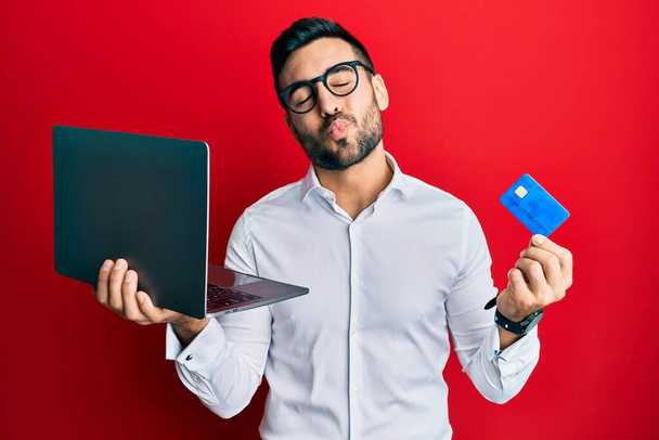 Young hispanic man wearing business style holding laptop and credit card looking at the camera blowing a kiss being lovely and sexy. love expression.  - Foto, Imagen