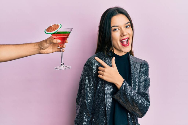 Young girl wearing sexy party jacket and hispanic arm offering cocktail smiling cheerful pointing with hand and finger up to the side  - Фото, изображение