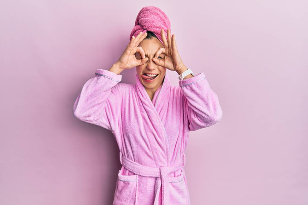 Young blonde woman wearing shower towel cap and bathrobe doing ok gesture like binoculars sticking tongue out, eyes looking through fingers. crazy expression.  - Fotografie, Obrázek