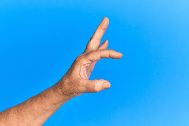 Mano de hombre hispano mayor sobre fondo azul aislado recogiendo y tomando lo invisible, sosteniendo el objeto con los dedos mostrando espacio  - Foto, Imagen