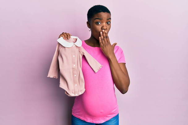 Jovem afro-americana esperando um bebê segurando roupas cobrindo a boca com a mão, chocada e com medo de erros. expressão surpreendida  - Foto, Imagem