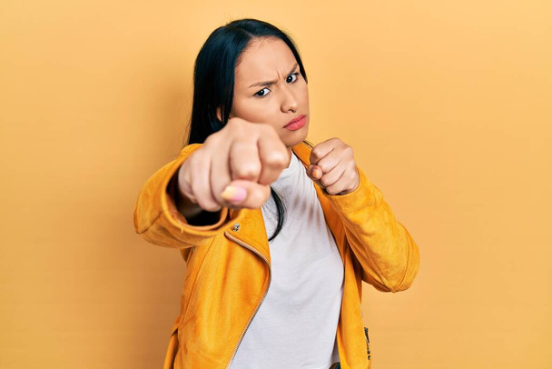 Hermosa mujer hispana con piercing en la nariz con chaqueta de cuero amarillo golpeando puño para luchar, ataque agresivo y enojado, amenaza y violencia  - Foto, Imagen