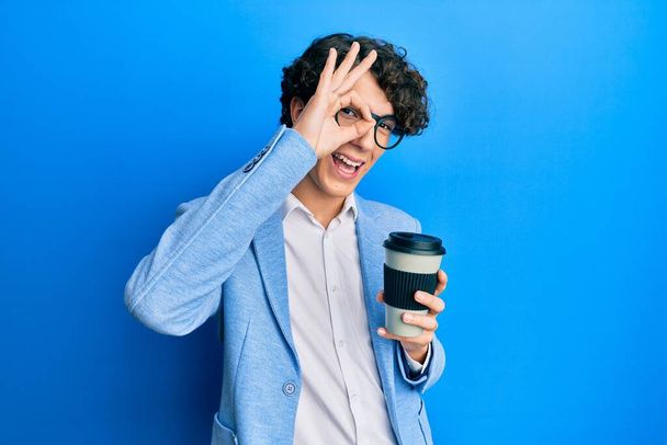 Ισπανόφωνος νεαρός άνδρας που φοράει σακάκι πίνοντας παίρνει τον καφέ χαμογελώντας χαρούμενος κάνει εντάξει σημάδι με το χέρι στο μάτι κοιτάζοντας μέσα από τα δάχτυλα  - Φωτογραφία, εικόνα