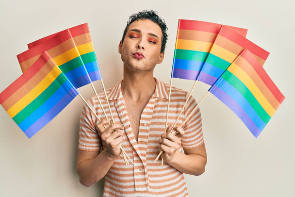 Hombre guapo usando maquillaje sosteniendo banderas de arco iris lgbtq mirando a la cámara soplando un beso siendo encantador y sexy. expresión de amor.  - Foto, Imagen