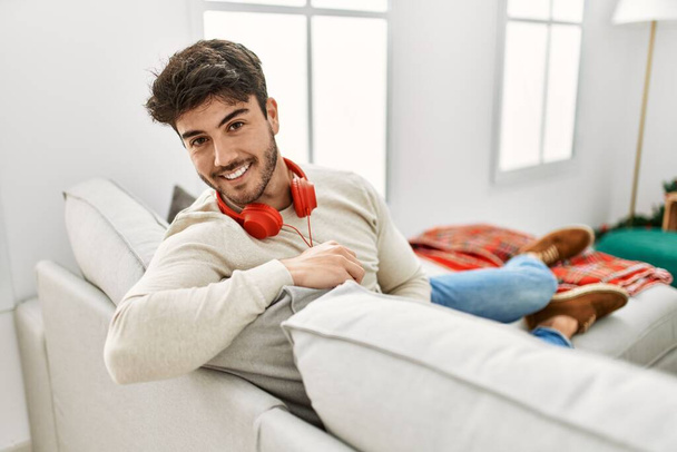 Joven hombre hispano celebrando la Navidad usando auriculares sentados en el sofá en casa. - Foto, imagen
