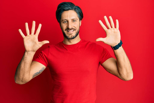 Joven hombre hispano vistiendo casual camiseta roja mostrando y señalando con los dedos número diez mientras sonríe confiado y feliz.  - Foto, Imagen