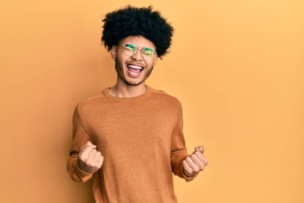 Young african american man with afro hair wearing casual winter sweater excited for success with arms raised and eyes closed celebrating victory smiling. winner concept.  - Photo, Image