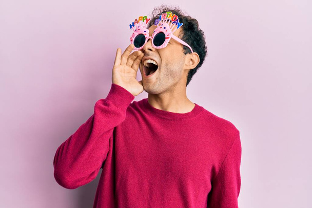 Hombre hispano guapo usando gafas de cumpleaños felices graciosas gritando y gritando fuerte a un lado con la mano en la boca. concepto de comunicación.  - Foto, imagen