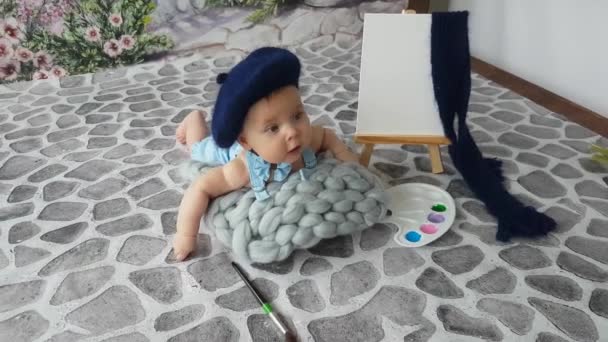 Un niño artista de cinco meses de edad en una boina azul está acostado en un pavimento con una paleta y caballete. Él realmente quiere dibujar, pero todavía no puede - Metraje, vídeo