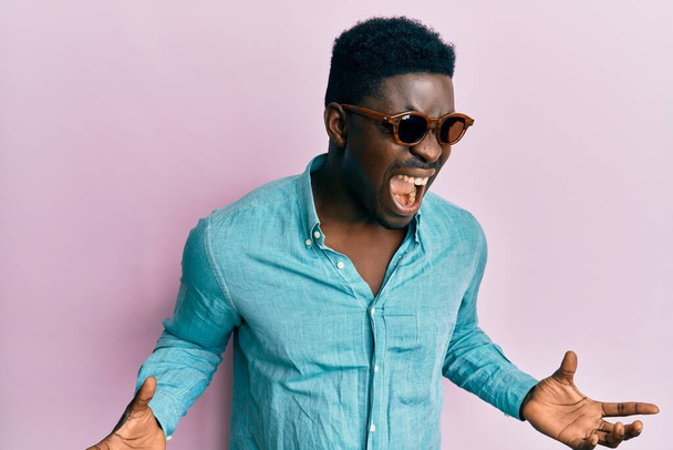 Handsome business black man wearing stylish sunglasses crazy and mad shouting and yelling with aggressive expression and arms raised. frustration concept.  - Photo, Image