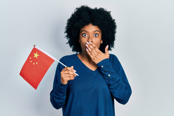 Fiatal afro-amerikai nő kínai zászlóval a szájában, kézzel, sokkolva és félve a hibáktól. meglepett kifejezés  - Fotó, kép