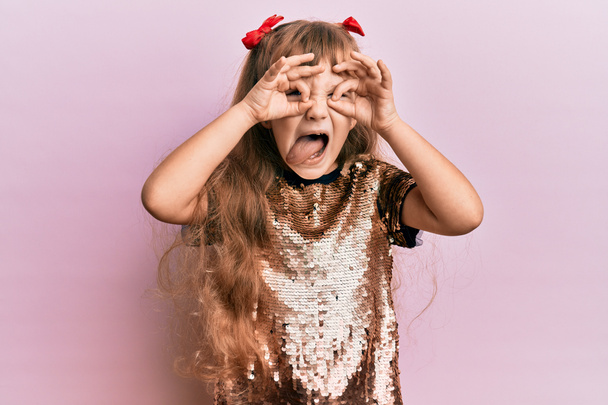 Kleine kaukasische Mädchen in festlichem Paillettenkleid machen eine gute Geste wie ein Fernglas, das die Zunge herausstreckt, die Augen blicken durch die Finger. Verrückter Ausdruck.  - Foto, Bild