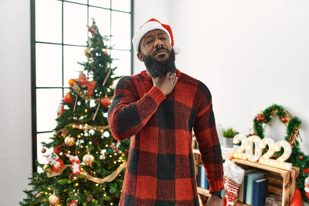 Αφροαμερικάνος που φοράει καπέλο Σάντα Κλάους στέκεται δίπλα στο χριστουγεννιάτικο δέντρο αγγίζοντας τον επώδυνο λαιμό, πονόλαιμο για γρίπη, θρόμβο και λοίμωξη  - Φωτογραφία, εικόνα