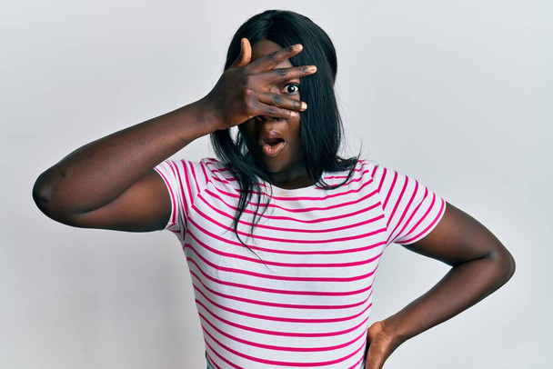 Mujer joven africana vistiendo camiseta de rayas casuales asomándose en shock cubriendo la cara y los ojos con la mano, mirando a través de los dedos asustados  - Foto, imagen
