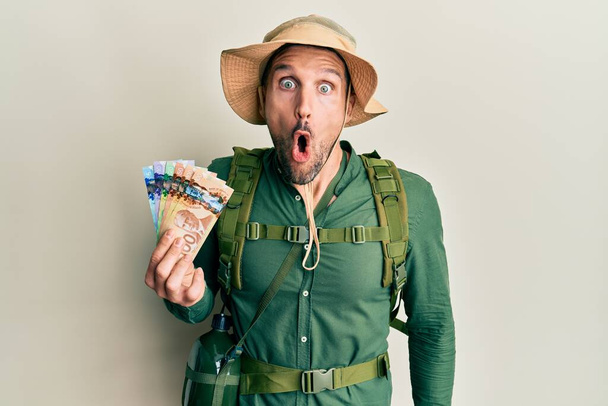 Όμορφος άνδρας με γενειάδα φορώντας καπέλο εξερευνητή κρατώντας καναδικά δολάρια φοβισμένος και έκπληκτος με ανοιχτό το στόμα για έκπληξη, δυσπιστία πρόσωπο  - Φωτογραφία, εικόνα