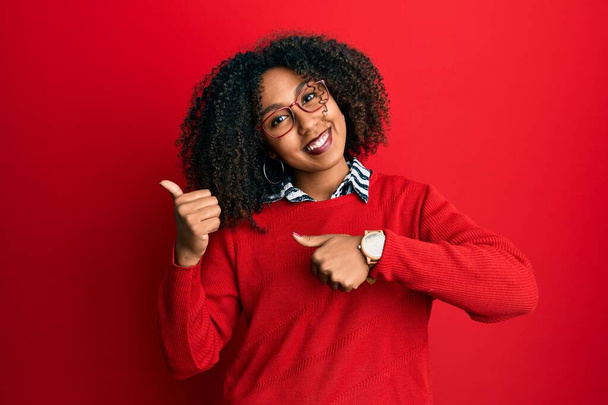 Gyönyörű afro-amerikai nő afro haj visel pulóver és szemüveg mutatva a háta mögött a kezét és hüvelykujját fel, mosolygós magabiztos  - Fotó, kép