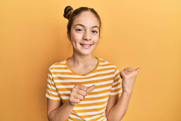 Krásná brunetka holčička na sobě ležérní pruhované tričko ukazuje na zadní straně s rukou a palci nahoru, s úsměvem sebevědomě  - Fotografie, Obrázek